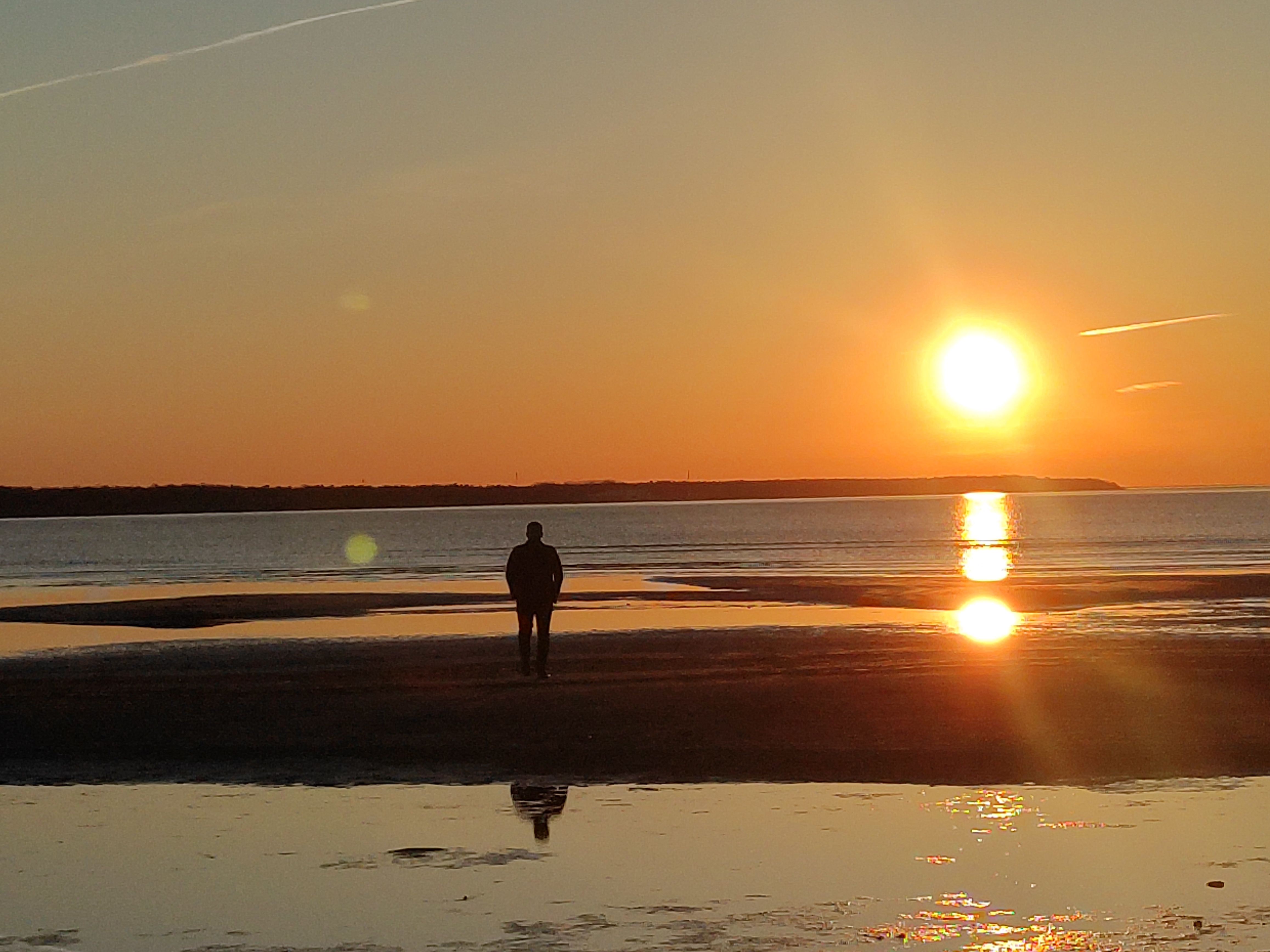A man walking towards us from the shoreline, with a beautiful sunset in the background